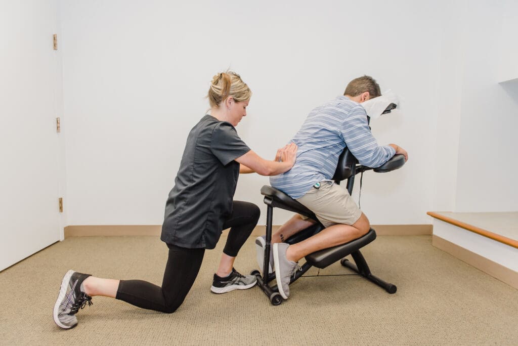 Corporate Chair Massages' owner Jeannette Sweeney kneels behind a male customer seated facing forward in a massage chair as she compresses his low back with both hands