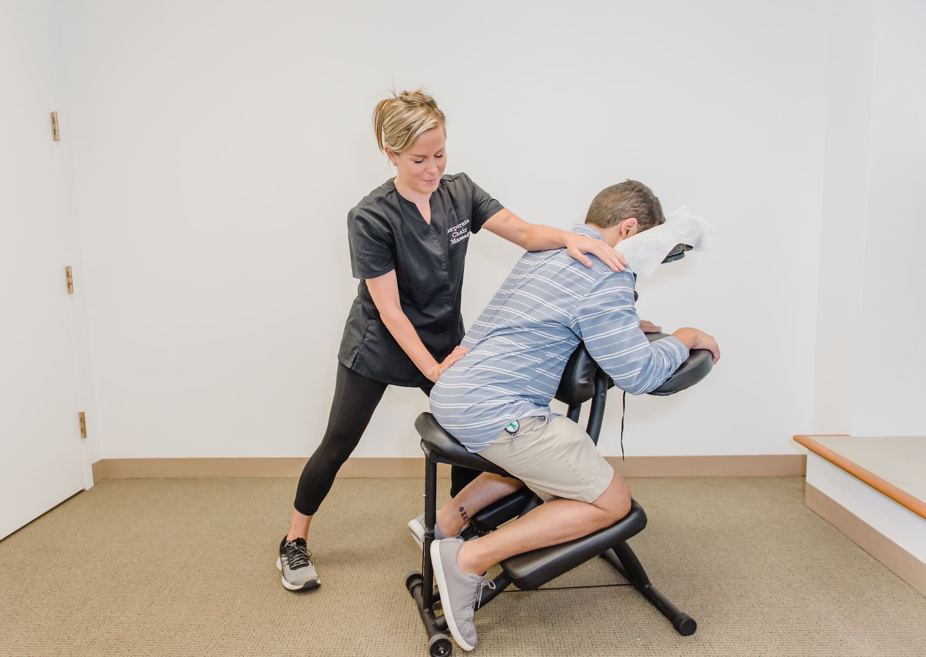 Corporate Chair Massages' owner Jeannette Sweeney provides a chair massage to a male customer. She uses her left arm to compress the upper back while the right arm gently applies pressure to the lower left hip creating a cross stretch for the spine.